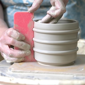 May include: A potter's hands shaping a light grey ceramic pot with a pink rib tool. The pot has a cylindrical shape with three horizontal bands. The potter's hands are covered in wet clay, indicating the pottery process.
