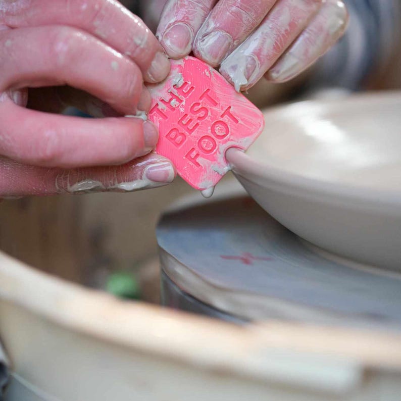 May include: A pink rubber stamp with the text "The Best Foot" being pressed onto a white ceramic plate.