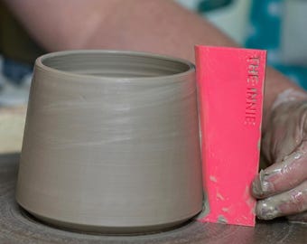 May include: A gray ceramic pot being shaped on a pottery wheel with a pink plastic tool labeled "The Innie" used to smooth the clay.