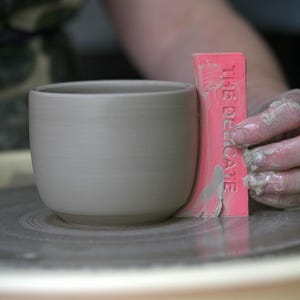 May include: A light grey ceramic cup is being shaped on a pottery wheel. A pink tool with the text "THE DELICATE" is used to smooth the cup's surface. The cup has a rounded shape and a flat base.