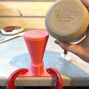 May include: A pink ceramic mold with a funnel shape is clamped to a surface with red clamps. A ceramic bowl is held in the background. The bowl has the text "Rhiannon Sparks" stamped on the bottom.