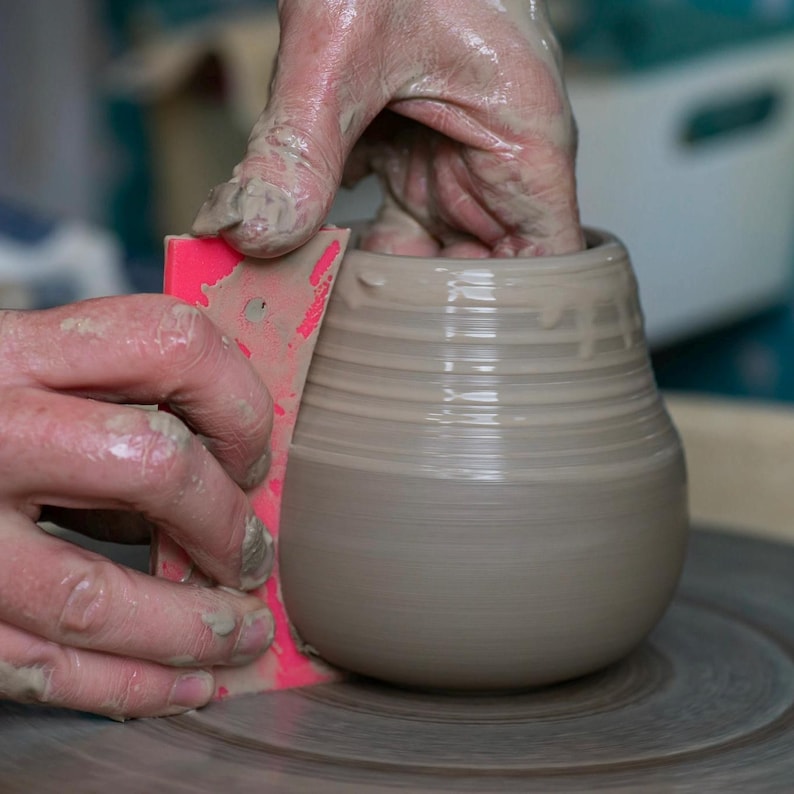 May include: A person's hands are shaping a gray clay pot on a pottery wheel using a pink tool. The pot has a rounded bottom and a wide opening.