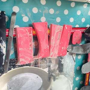 May include: Four pink pottery tools with the words "The Straight", "The Curvy", "The Irrie", and "The Foutie" printed on them. The tools are arranged on a table with a pottery wheel in the background.