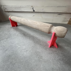 May include: A wooden rolling pin with rounded ends is resting on two bright pink triangular stands. The rolling pin is light brown and the stands are a vibrant pink.
