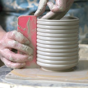 May include: Hands shaping a white ceramic vase with horizontal ridges. A pink rib tool is used to refine the clay. The hands are covered in wet clay, showing the pottery process. The vase is on a wooden surface.
