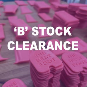 May include: Pink plastic shoe inserts, some stacked, some scattered, with the text "B STOCK CLEARANCE". The inserts have the words "THE BEST FOOT" printed on them. The background is a wooden surface.