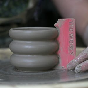 May include: A gray, unfinished ceramic pot with a rounded, stacked design. A pink tool with the text "THE WIGGLY" is used to shape the pot. The pot is on a gray surface, likely a pottery wheel.