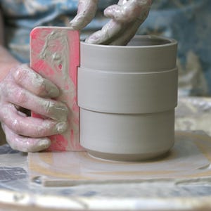 May include: A potter's hands shaping a light gray ceramic mug with a pink ribbing tool. The mug has a unique design with a band around the middle. The potter's hands are covered in wet clay, highlighting the handmade process.