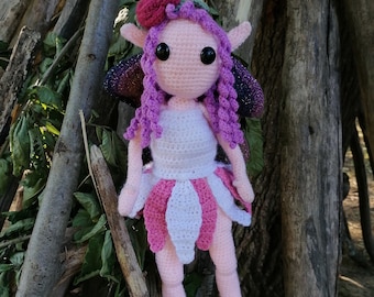 May include: A crocheted fairy doll with pink and white clothing, purple hair, and sparkly wings. The doll is hanging from a branch.