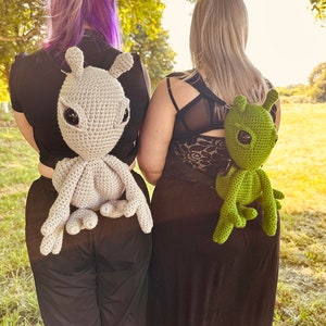 May include: Two people wearing black clothing with crocheted alien backpack toys. The alien on the left is gray and the alien on the right is green.