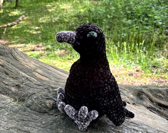May include: A black crocheted crow with a grey beak and large black eyes. The crow is sitting on a grey log.
