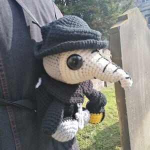 May include: A handmade crochet Plague Doctor doll, featuring a black hat, mask, and cloak. The doll has a long, white beak and large black eyes. It is holding a yellow and black lantern. The doll is attached to a black strap.