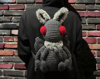 Könnte beinhalten: Ein grauer Häkel-Fledermaus-Rucksack mit roten Augen. Der Rucksack wird von einer Person getragen, die einen schwarzen Hoodie trägt.