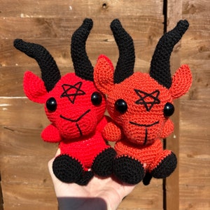 May include: Two red crocheted plush toys shaped like a goat with black horns and black feet. Each toy has a black pentagram on its forehead and black eyes.