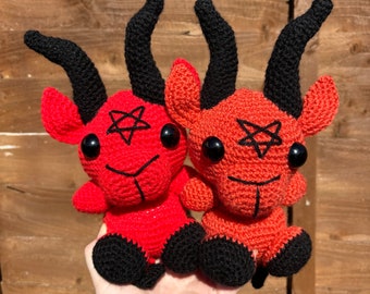Puede incluir: Dos juguetes de peluche de crochet rojo con forma de cabra con cuernos negros y patas negras. Cada juguete tiene un pentagrama negro en la frente y ojos negros.