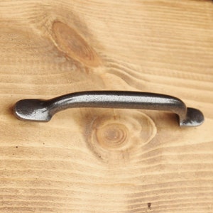 May include: A silver-toned metal drawer pull with a curved design. The pull is mounted on a light brown wooden surface.