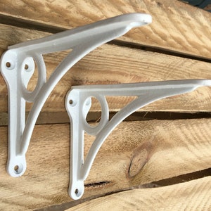 May include: Two white metal shelf brackets with a decorative design. The brackets are made of cast iron and have a white finish.