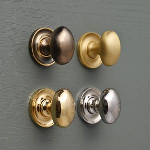 May include: Four cabinet knobs in different finishes: antique brass, brushed gold, polished gold, and polished nickel. Each knob has a round backplate and an oval-shaped handle. The knobs are mounted on a gray surface.