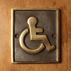 May include: A rectangular brass handicap accessible sign with a gold-colored wheelchair symbol on a dark background. The sign is mounted on a wooden surface and has two screw holes.