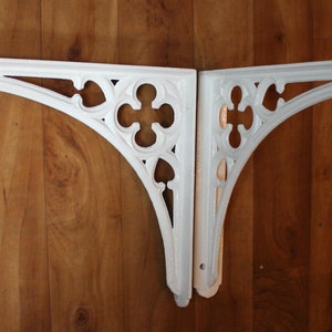 Puede incluir: Dos soportes de estante de hierro fundido blanco decorativos con un dise&ntilde;o g&oacute;tico. Los soportes est&aacute;n montados en una pared de madera.