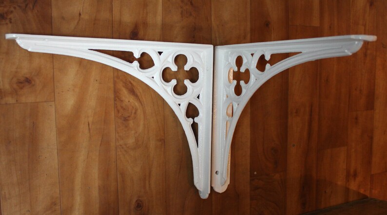 Puede incluir: Dos soportes de estante de hierro fundido blanco decorativos con un dise&ntilde;o g&oacute;tico. Los soportes est&aacute;n montados en una pared de madera.