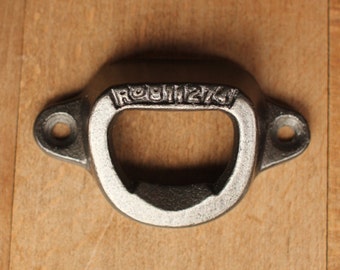 May include: A black metal bottle opener with a rounded shape and a small hole on each side. The opener has the text "ROSHMZ" stamped on the top.