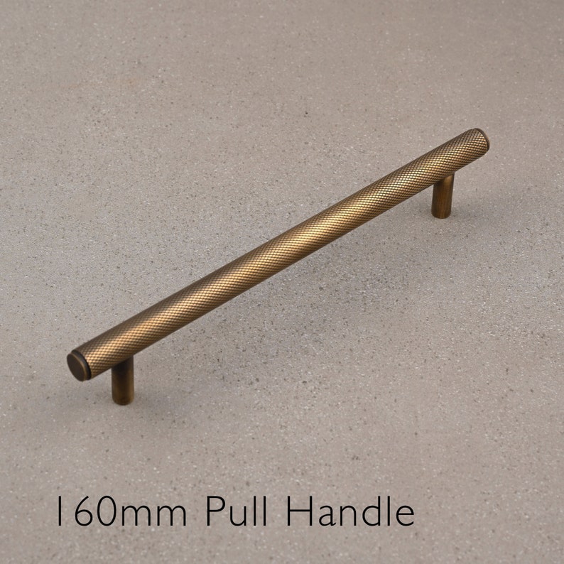 May include: A 160mm long, gold-toned metal pull handle with a textured surface. The handle is designed for use on cabinets, drawers, or doors.