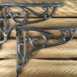 May include: Two dark gray cast iron shelf brackets with ornate scrollwork and a triangular shape. The brackets are mounted on a wooden surface. Each bracket has a small text stamp that reads "4 07".