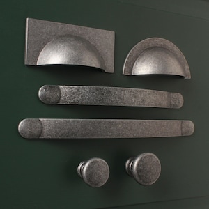 May include: A set of five silver-toned cabinet hardware pieces, including two knobs, two pulls, and one cup pull. The hardware has a distressed finish and is displayed against a dark green background.