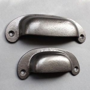 Könnte beinhalten: Zwei schwarze Metall-Schubladengriffe mit abgerundeter, leicht geschwungener Form. Die Griffe haben eine glatte, leicht strukturierte Oberfläche.