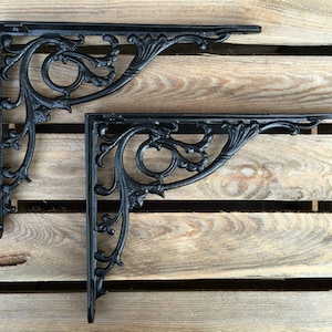 May include: Two black cast iron shelf brackets with a decorative scroll design. The brackets are shown against a wood background.