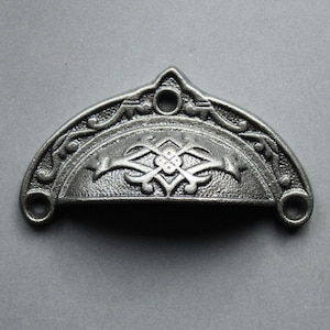 May include: A silver cast iron drawer pull with an ornate design. The pull is shaped like a half-circle with a decorative floral pattern in the center.