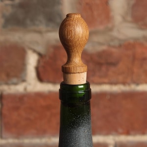 May include: A wooden bottle stopper with a rounded top and a cork base. The stopper is a light brown color and has a natural wood grain.