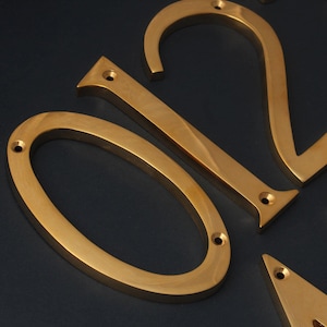 K&ouml;nnte beinhalten: Goldfarbene Hausnummern aus Metall, 0, 1 und 2, mit geb&uuml;rsteter Oberfl&auml;che. Die Zahlen sind auf einer dunkelblauen Oberfl&auml;che befestigt.