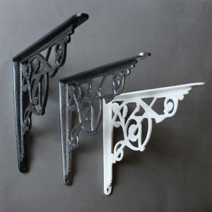 Puede incluir: Dos soportes decorativos para estantes, uno negro y otro blanco, con un diseño floral ornamentado. Los soportes son de metal y tienen un estilo vintage.