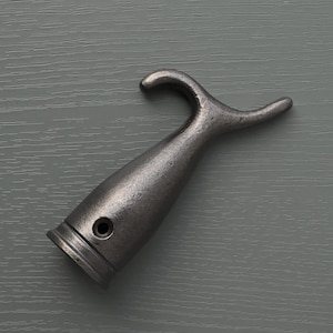 Puede incluir: Un gancho de metal gris oscuro envejecido, con una parte superior curvada y una base cilíndrica. El gancho tiene una superficie texturizada y un pequeño agujero cerca de la base. El diseño recuerda a la ferretería náutica.