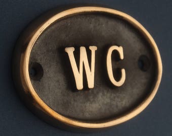 Op de afbeelding: Een zwart ovaalvormig bord met een gouden rand. Het bord zegt "WC" in gouden letters.