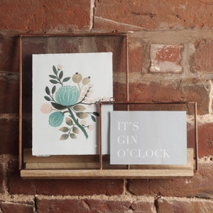 May include: Two copper framed glass picture frames on a wooden shelf against a brick wall. The larger frame holds a floral print with pink, green, and blue colours. The smaller frame holds a grey print with the text "It's Gin O'Clock".