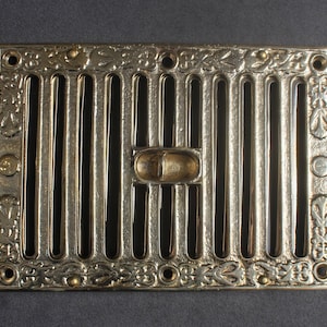 May include: A gold-toned metal vent cover with an ornate design and a rectangular shape. The vent cover has a series of vertical bars and a decorative center piece.