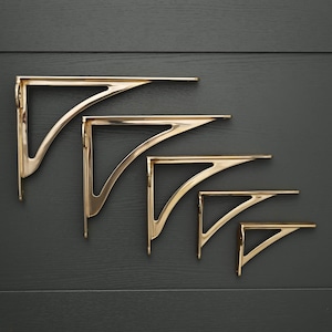 Puede incluir: Cinco soportes para estantes en forma de L hechos de metal pulido en tono dorado. Cada soporte presenta un refuerzo triangular con un recorte interior curvo. Los soportes están dispuestos en orden de tamaño decreciente, mostrando un diseño consistente en todas las piezas.