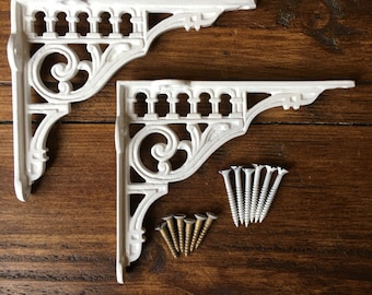 Puede incluir: Dos soportes de estante de hierro fundido blanco con volutas ornamentadas y un diseño de barandilla decorativo. Los soportes están sobre una superficie de madera con tornillos.