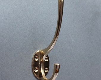 May include: A brass coat hook with a curved design and an oval base. The hook has four screw holes for mounting.