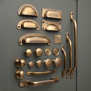 Può includere: Un assortimento di ferramenta per mobili in ottone antico, tra cui maniglie a tazza, pomelli e maniglie. L'hardware è disposto su una superficie grigia, mostrando varie forme e dimensioni per progetti di cucina o mobili.