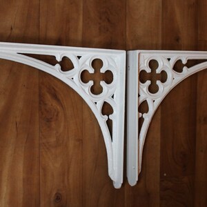 Puede incluir: Dos soportes de estante de hierro fundido blanco decorativos con un dise&ntilde;o g&oacute;tico. Los soportes tienen forma de arco apuntado con detalles intrincados.