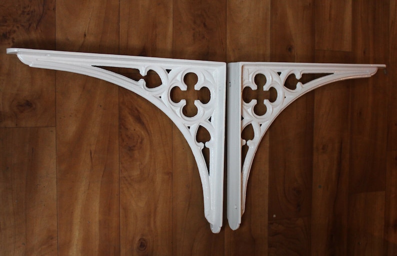 Puede incluir: Dos soportes de estante de hierro fundido blanco decorativos con un dise&ntilde;o g&oacute;tico. Los soportes tienen forma de arco apuntado con detalles intrincados.