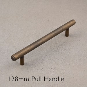 May include: A 128mm long, antique brass pull handle with a textured finish. The handle is shown on a light gray background.
