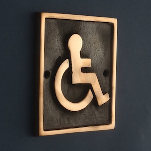 May include: A bronze plaque with a raised, gold-colored wheelchair accessibility symbol. The symbol is on a black background.