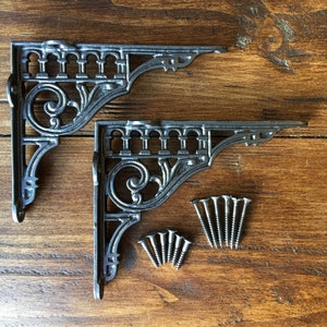 May include: Two ornate metal shelf brackets with a decorative scroll design. The brackets are made of cast iron and have a black finish. The brackets are shown on a wooden surface with screws.