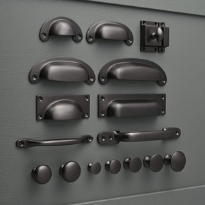 Op de afbeelding: Een verzameling zwarte kastbeslag, waaronder grepen, knoppen en een grendel. Het beslag is gerangschikt op een grijze achtergrond.