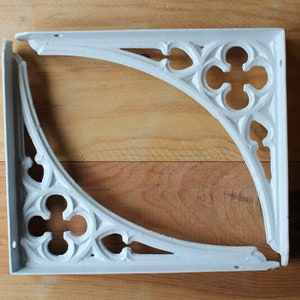 May include: Two white metal shelf brackets with a decorative, ornate design. The brackets are shaped like a right angle and have a pointed, Gothic-style arch.